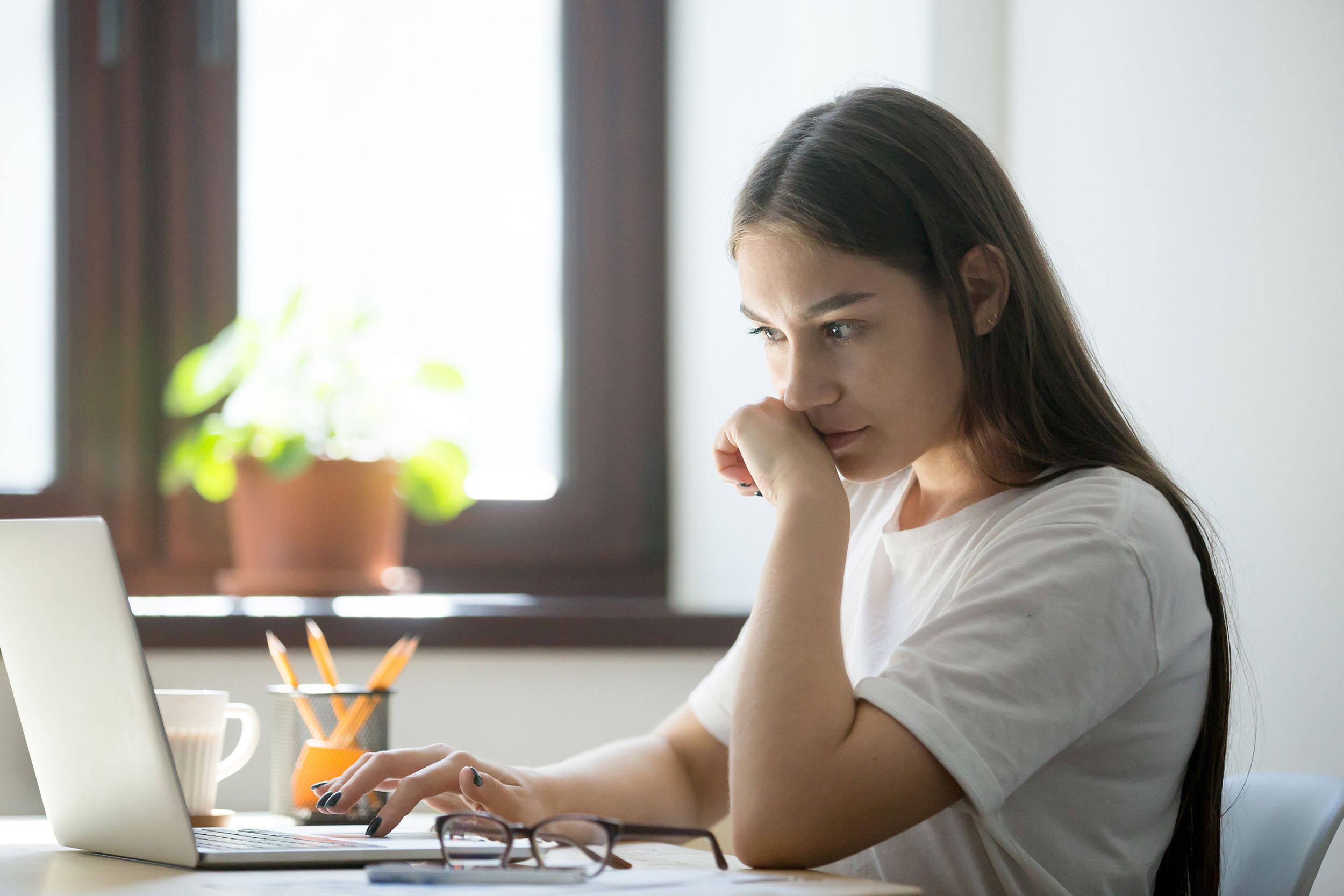 Side-view of an attentive woman sitting at her desk on a MacBook Air, light coming through a window, a potted plant on the window sill, blurred in the background