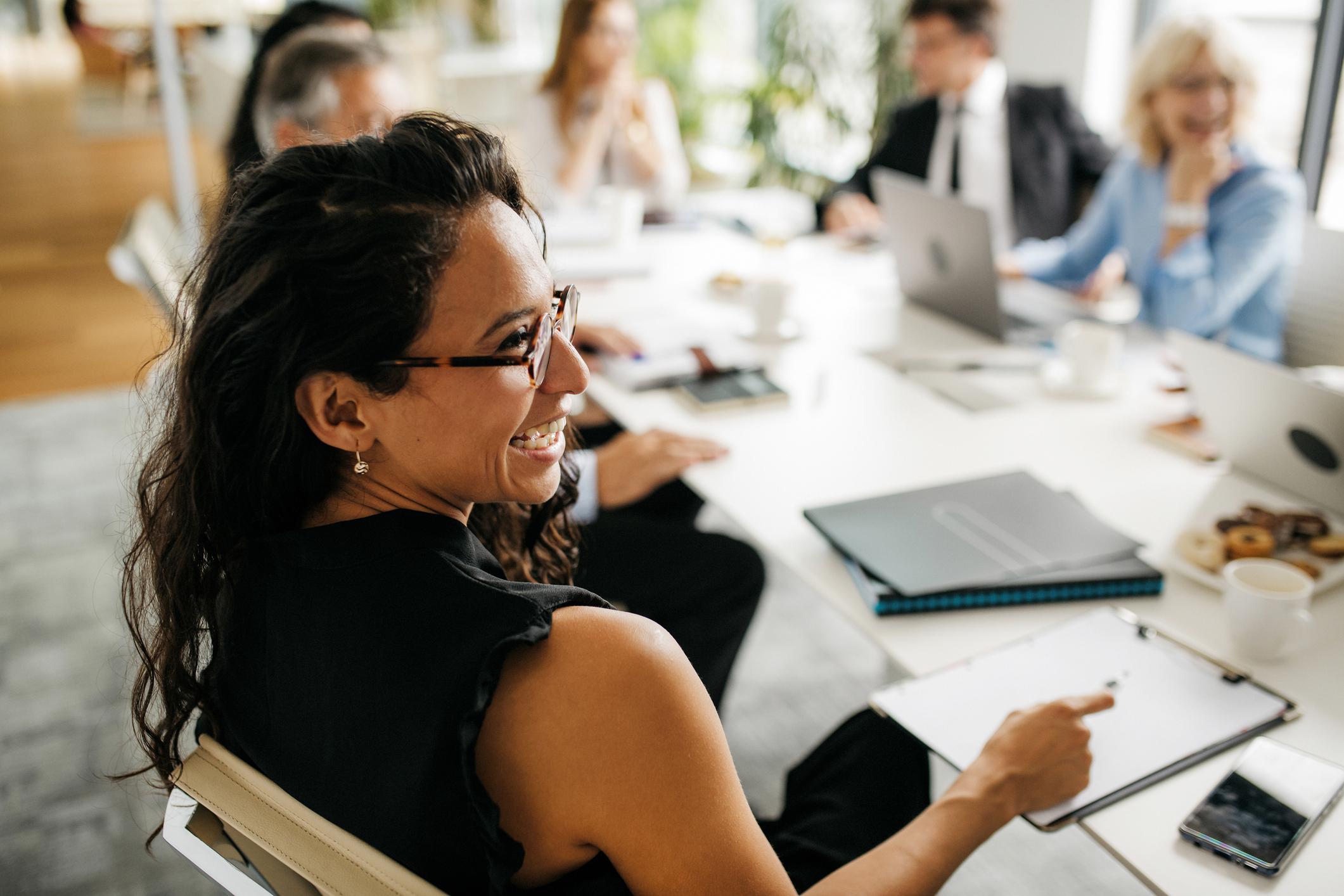 Woman sitting in a conference room table with several coworkers, smiling towards the right, selective focus, blurred background of coworkers and room