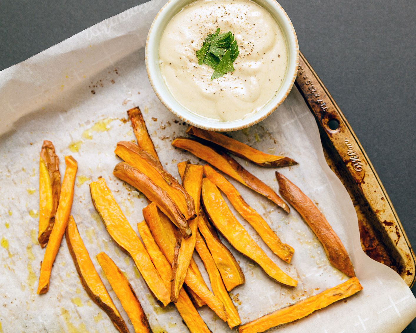 Sweet potato fries on a wooden tray on parchment paper with tahini garnished with fresh chopped herbs in a small white bowl