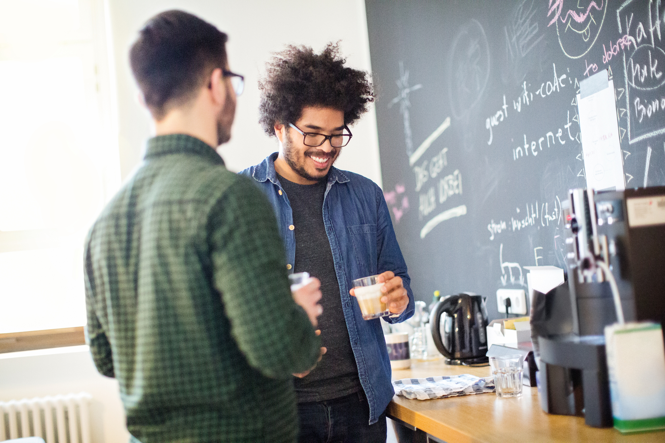 Two young male coworkers talking while having coffee in a startup office, sunlight coming through window and writing on chalkboard in the background