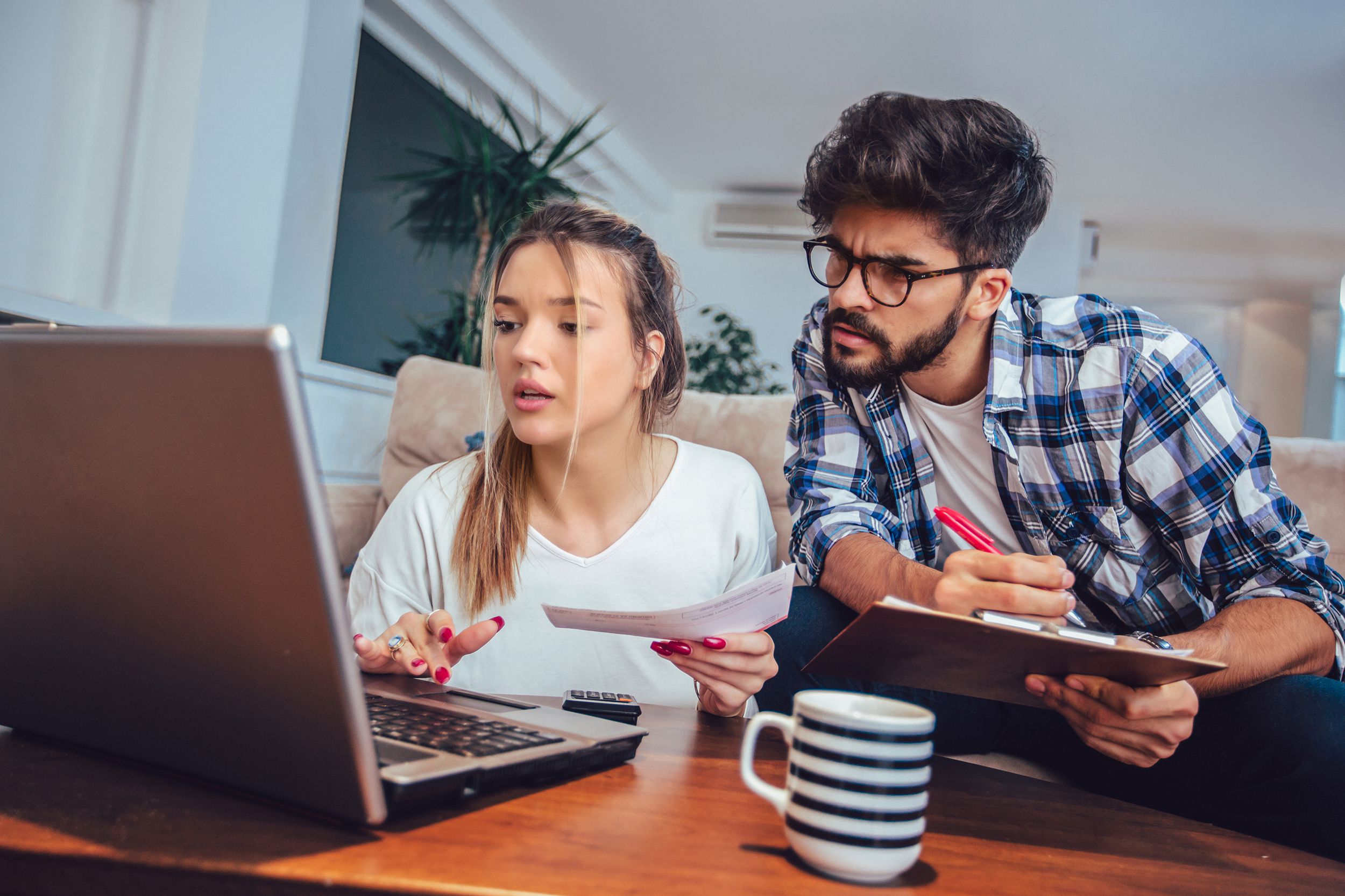 Young couple doing paperwork together in living room at home, woman is looking at laptop and man holding paper and pen, with closeup of coffee mug, on wooden table