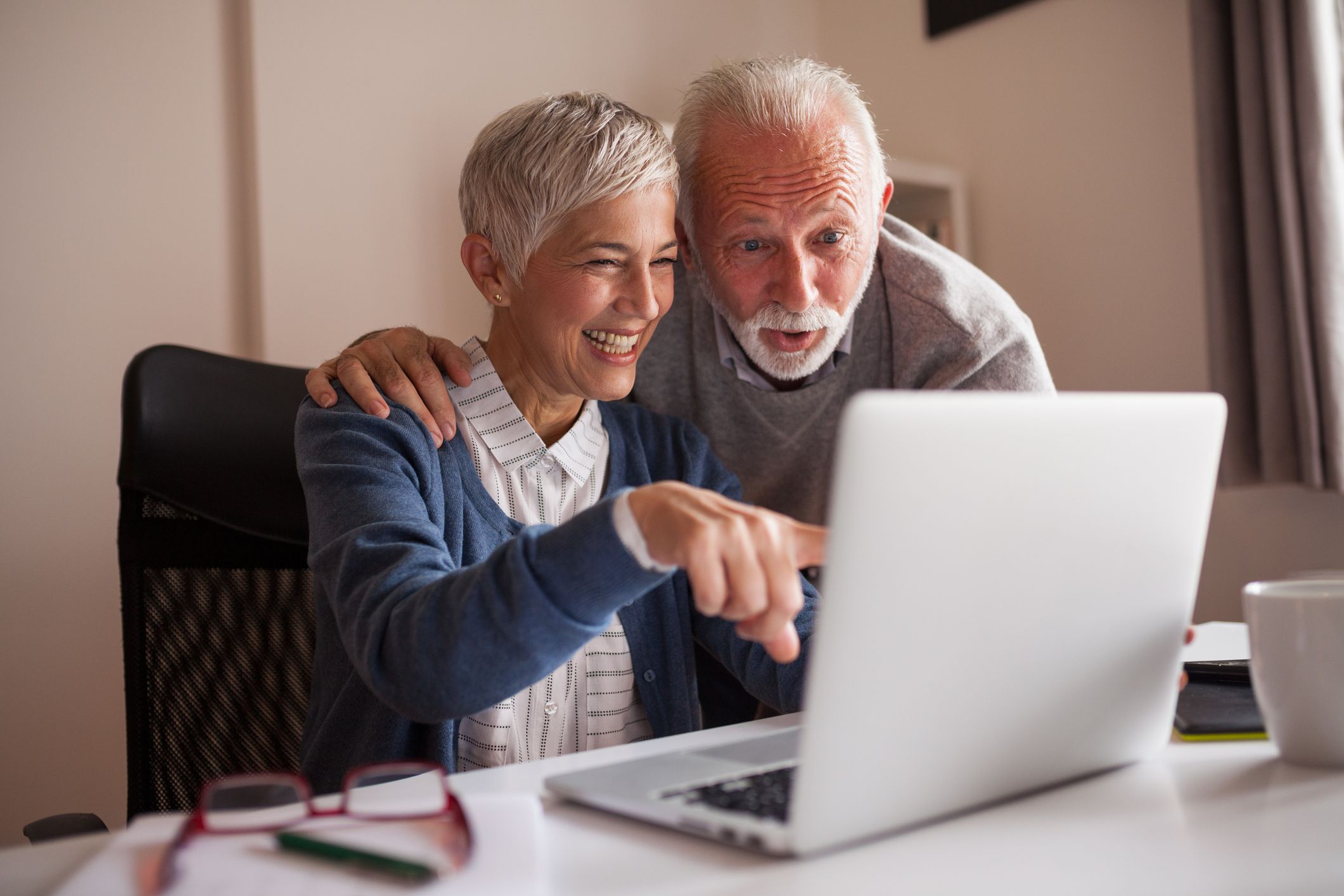 Youthful looking senior couple at laptop, woman is pointing to screen and man has surprise look on face, laptop on table at home