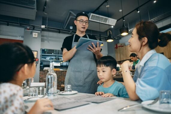 A restaurant server in a gray apron holds a tablet while taking an order from a family at a table.