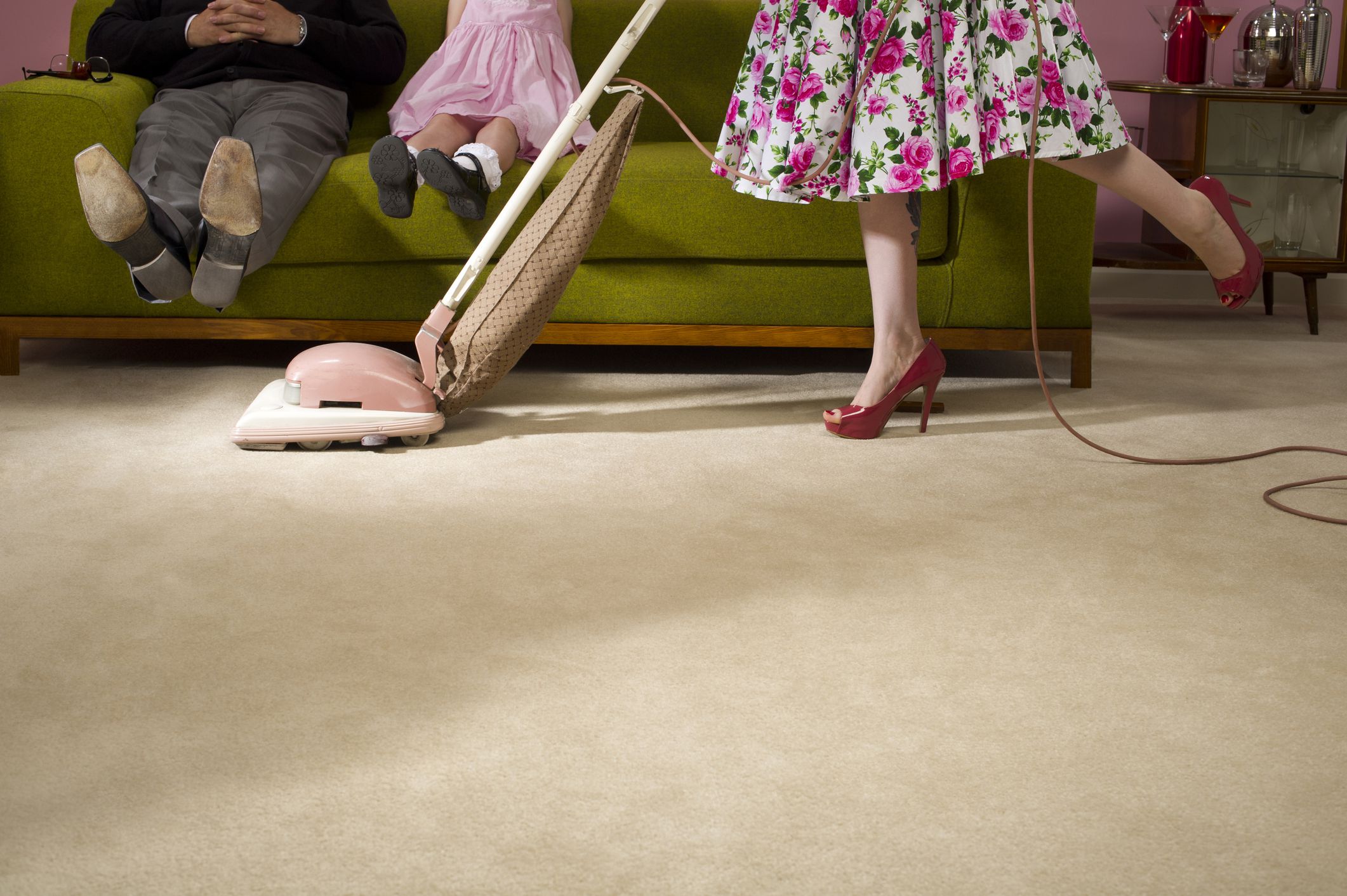 1950s home, child and man sitting on couch with legs up as woman in heels vacuums carpet, view of knees and below