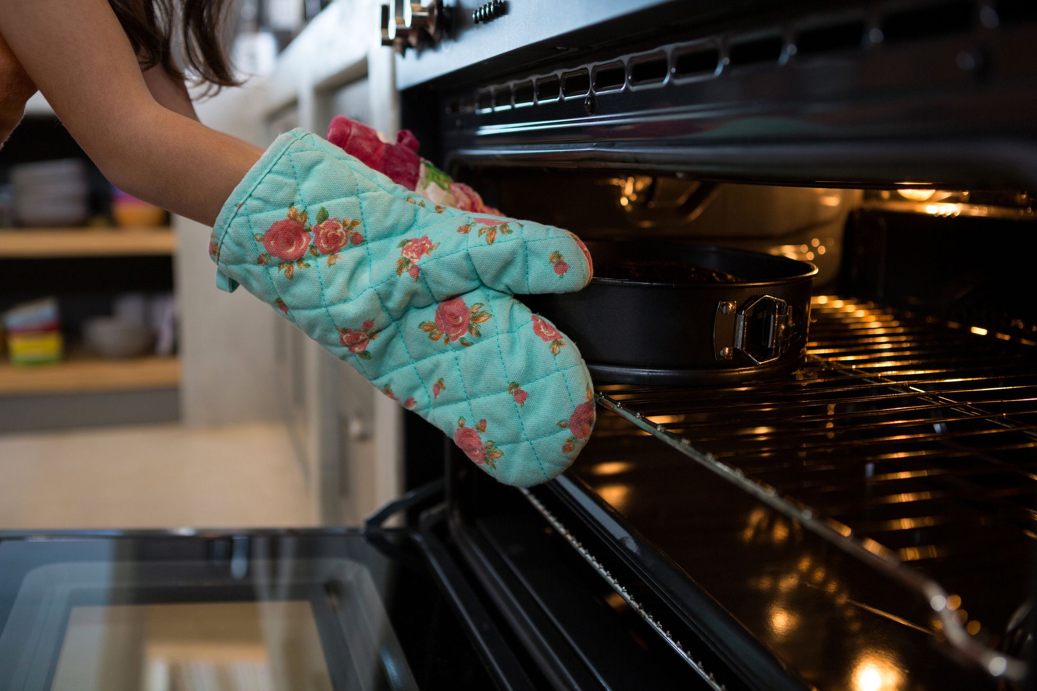 Cropped hands of girl wearing glove putting container with cake in oven at kitchen
