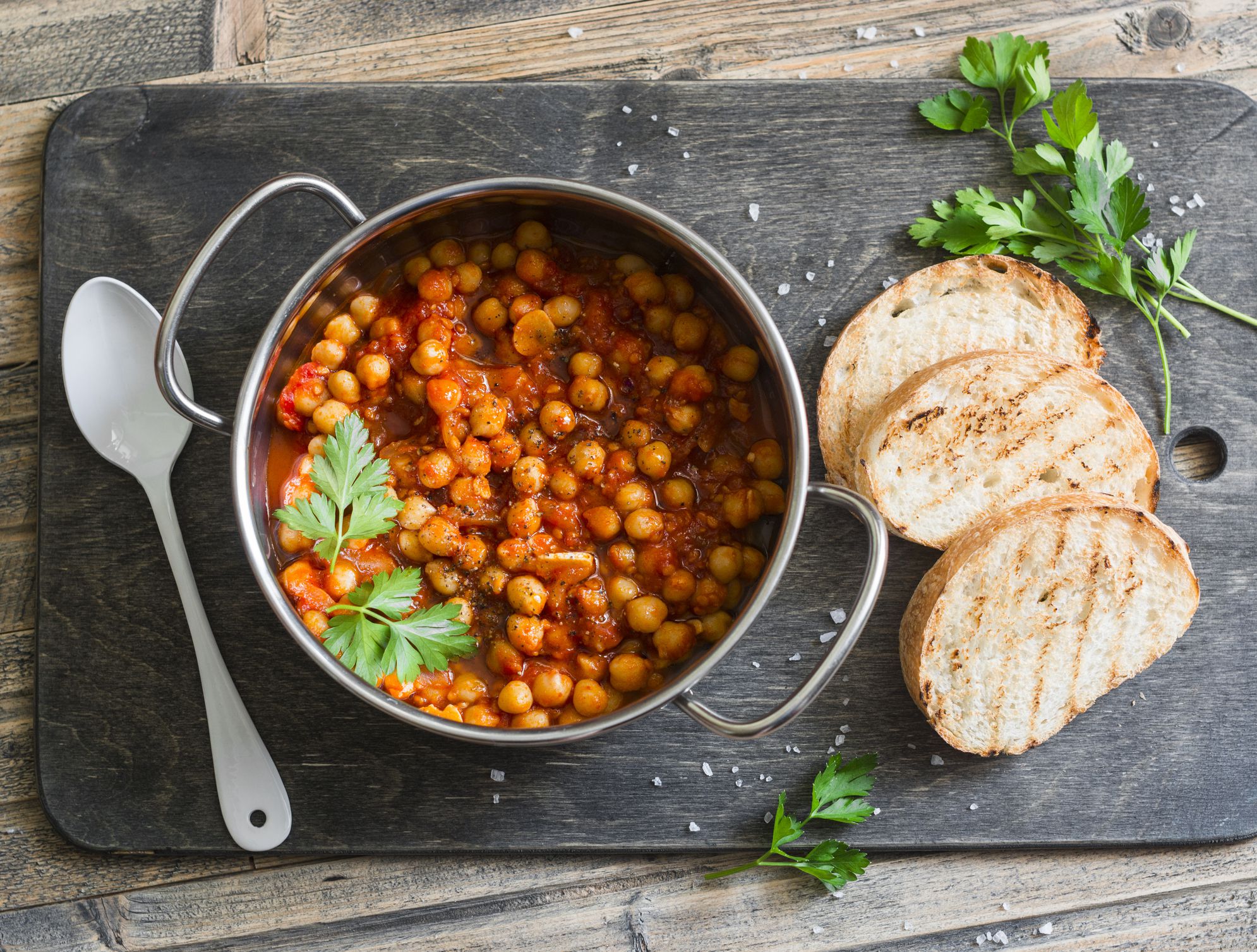 Tomato sauce braised chickpeas in a pot, and grilled bread. Delicious vegetarian lunch on a rustic wooden background, top view