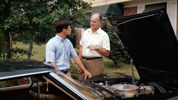 A young man and an older man stand next to a car with its hood open, having a conversation in a suburban yard with trees and a house in the background.