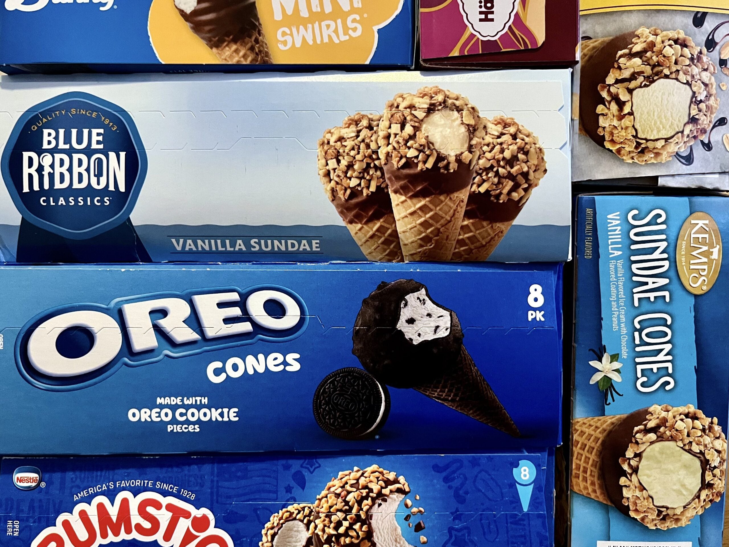 Boxes of various ice cream cones, including Blue Ribbon Classics Vanilla Sundae, Oreo Cones, and Kemps Sundae Cones, arranged closely together in a freezer.