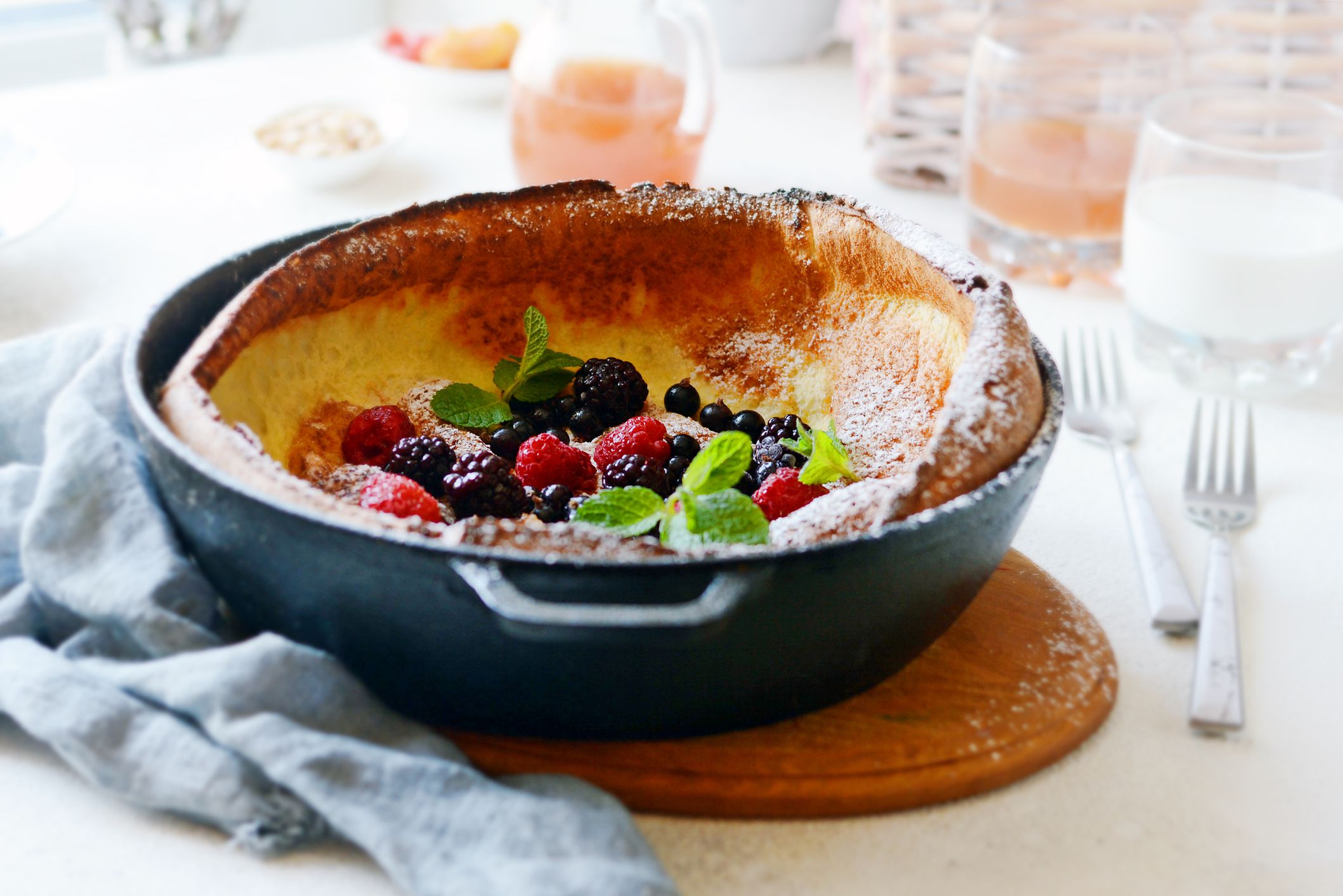 Fresh homemade dutch baby pancake with powdered sugar, fresh berries and mint for breakfast with a glass of milk and apple juice on a light background. Delicious breakfast in a sunny interior.