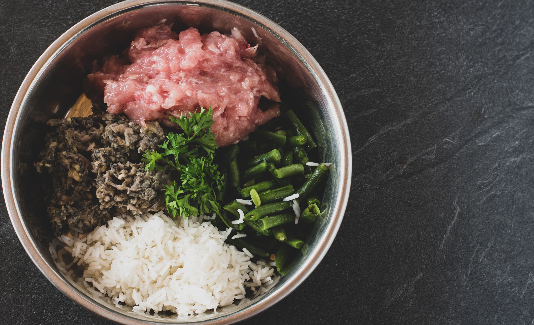 A bowl with fresh and raw dog food made with chicken meat and rumen served with cooked rice and green beans isolated on dark table background with copy space