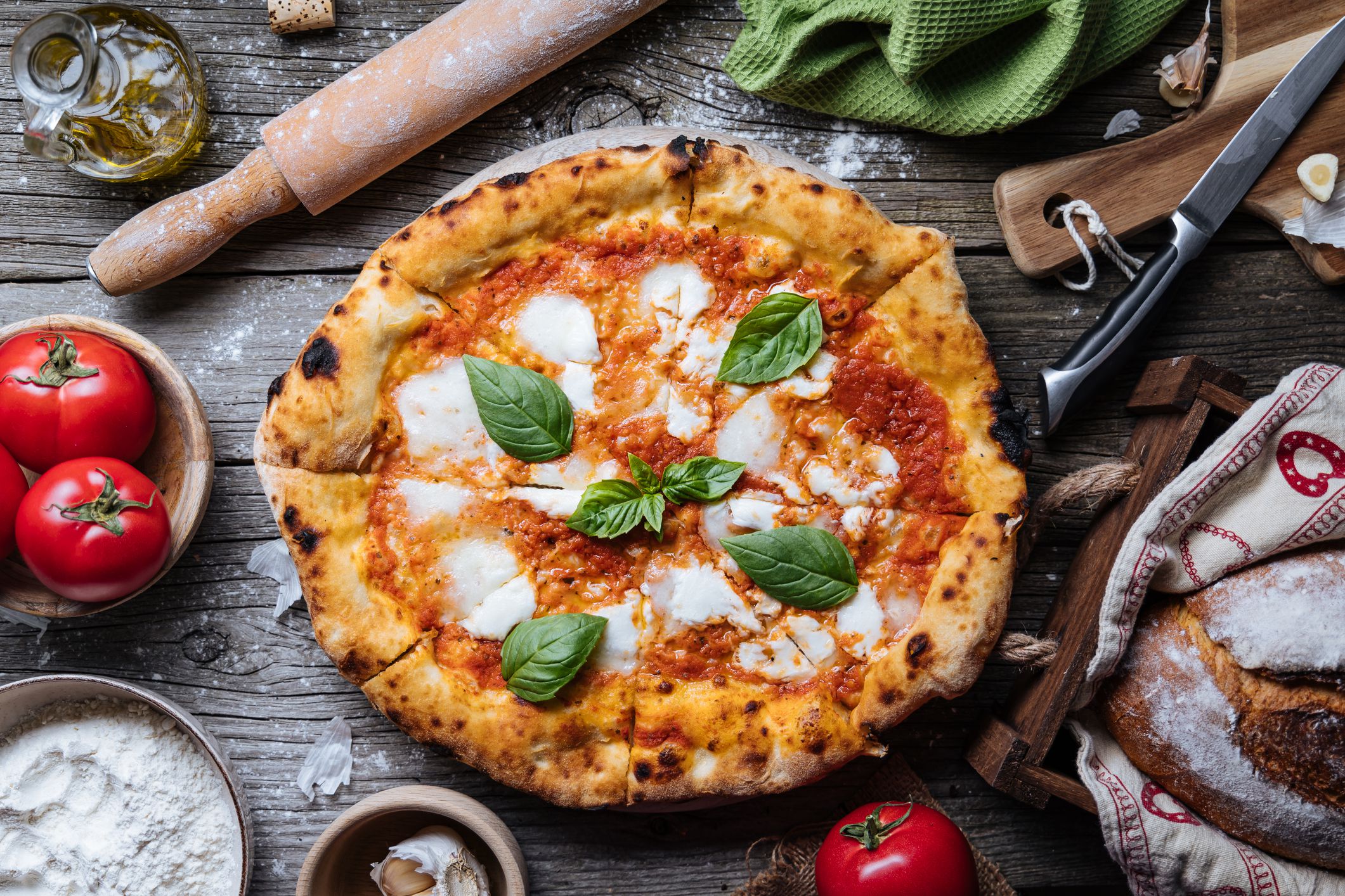 Italian Pizza Napoletana in Messy Kitchen with Cooking Ingredients such as tomato, olive oil, garlic, basil, rolling pin, flour, knife, cutting board