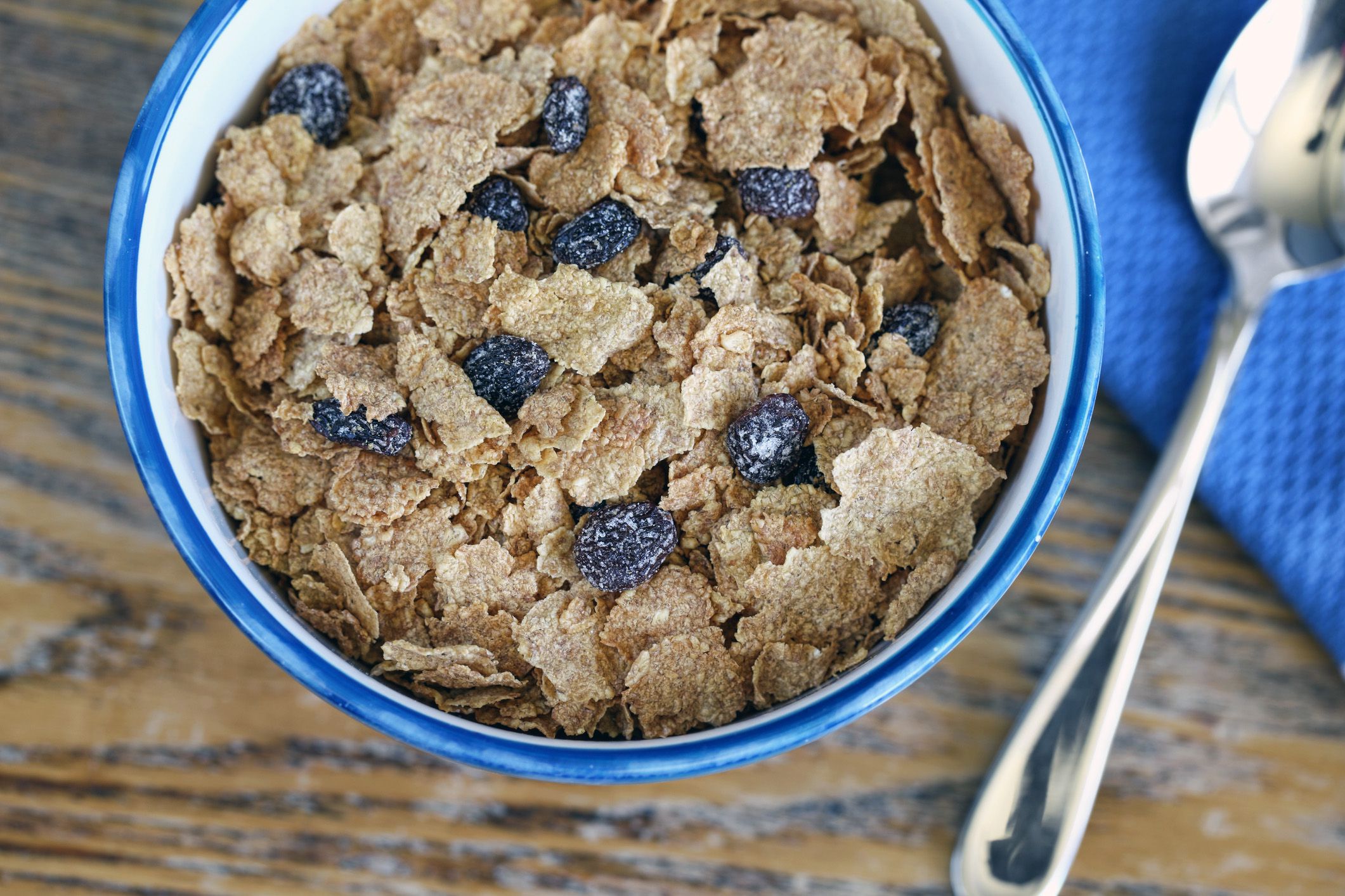 Bran cereal with raisins in a white bowl with a blue trim, a spoon, and blue napkin on a rustic wood table