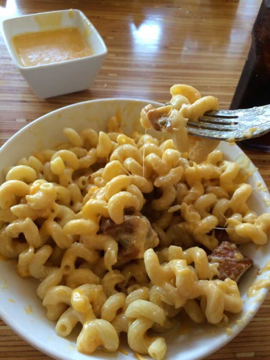 Closeup of Noodles & Company Wisconsin Mac & Cheese in a white bowl with a fork with a bite on the side of it, on a wooden table with cheese dipping sauce on the top side at Noodles & Company