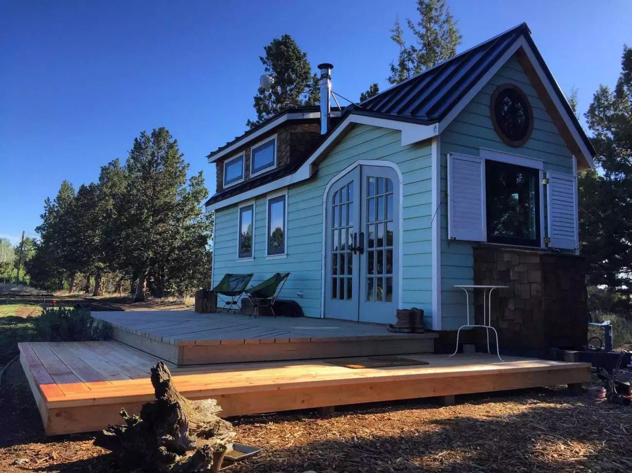 Front exterior of Mighty Small Homes The Tiny, painted light green with wooden and white accents, wooden two-step patio in the foreground, on a rustic lawn with many trees