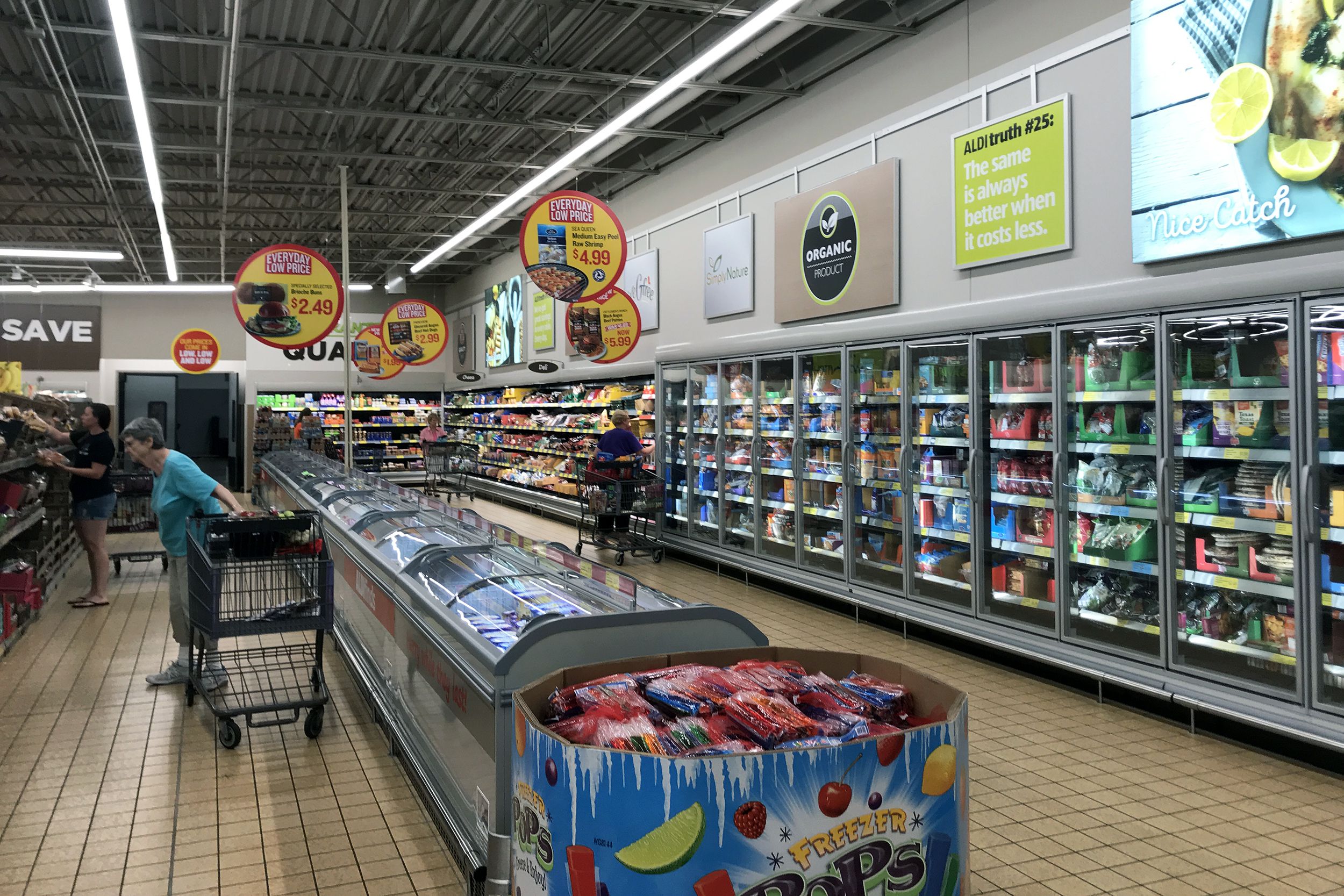 Frozen section of an Aldi store, wall of frozen goods with freezer in middle of a large aisle, clean store with three people with carts shopping