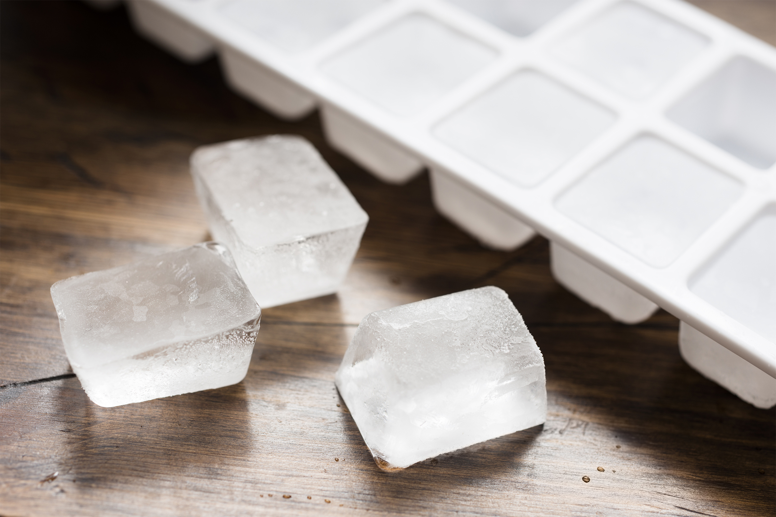 Ice cube tray filled with ice cubes and three loose ice cubes, selective focus of loose ice cubes, on a wooden table