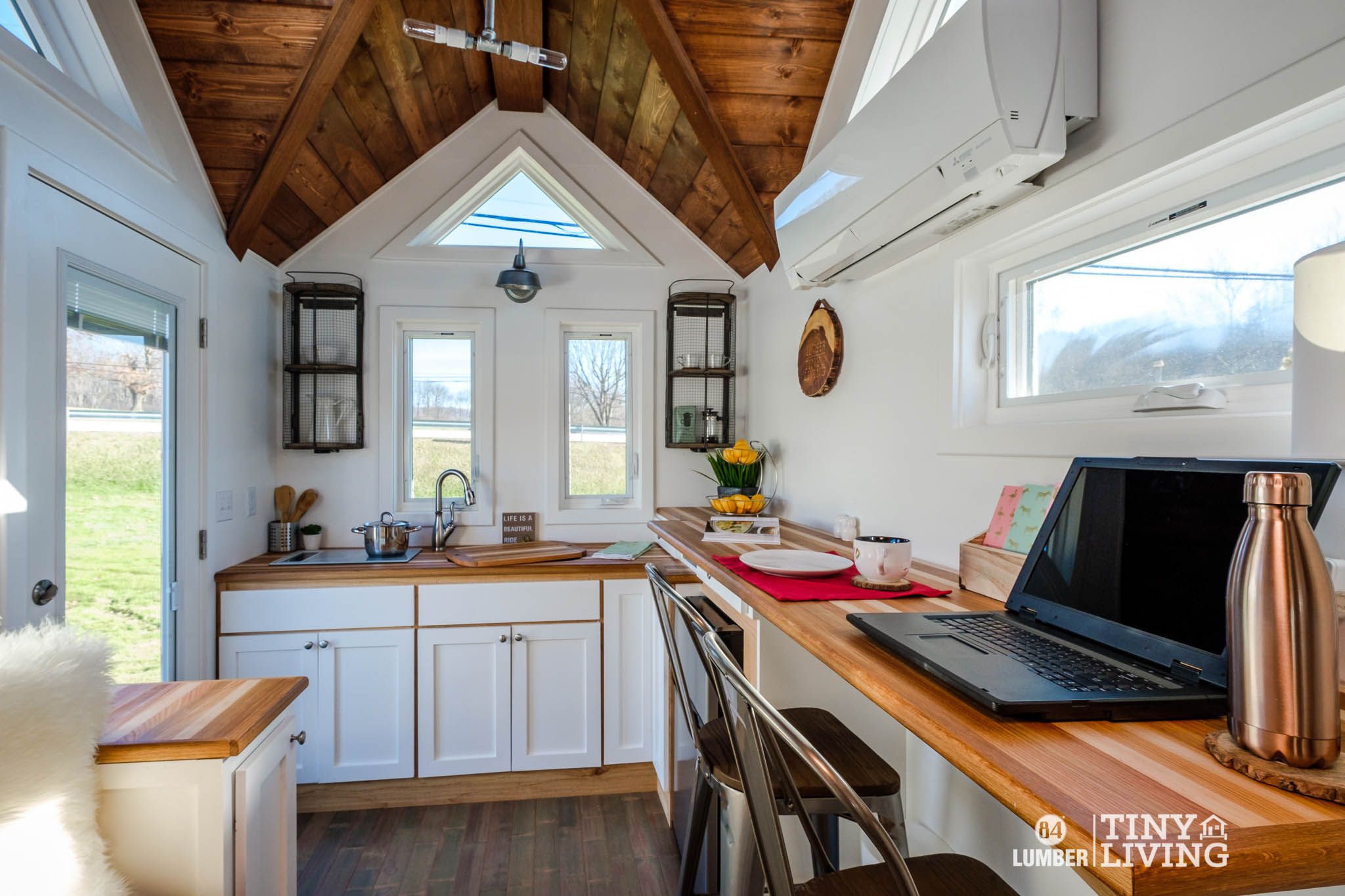 Interior of 84 Lumber Tiny Living Countryside Kit House, completed with lots of living accouterments, on a large piece of grass land as seen through the windows on a sunny day