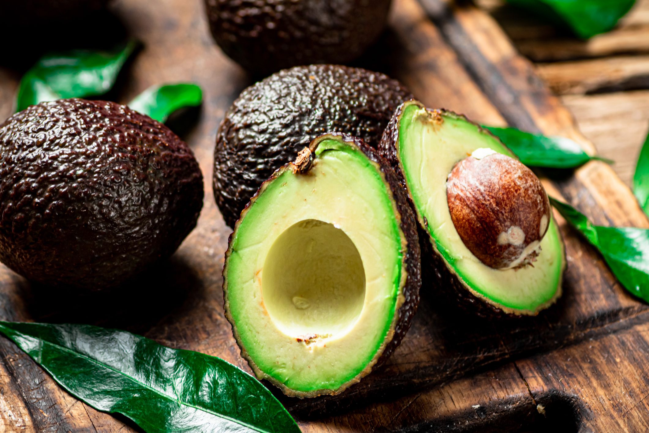 Closeup of two fresh cut halves of an avocado surrounded by several whole avocados on a wooden cutting board with a blurred background