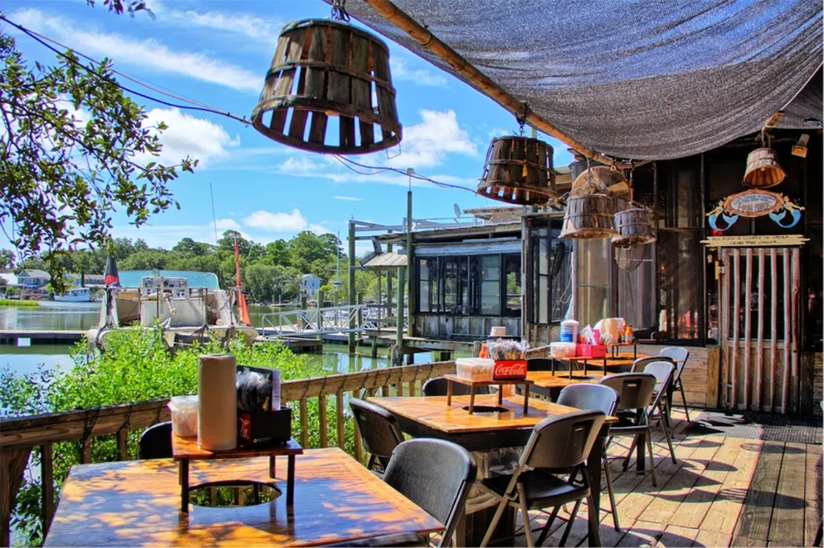 Outdoor restaurant patio with wooden tables and chairs overlooks a waterfront, offering one of the best dining experiences in America. Rustic basket lights and nautical decor create an inviting atmosphere under a shaded roof, with trees and boats in the background.