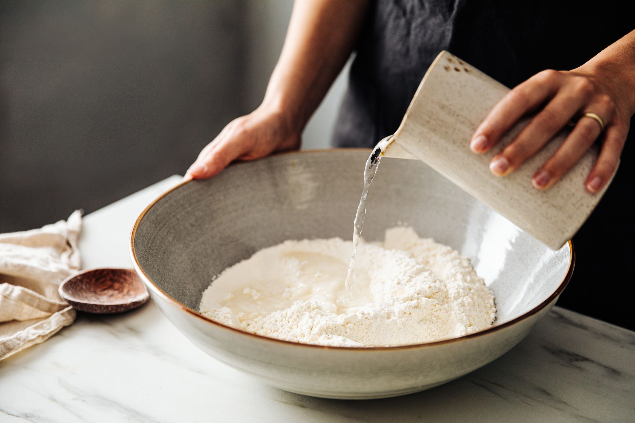 Midsection of woman pouring water on rye and wheat flour in bowl. Female is standing in kitchen. She is preparing sourdough bread.