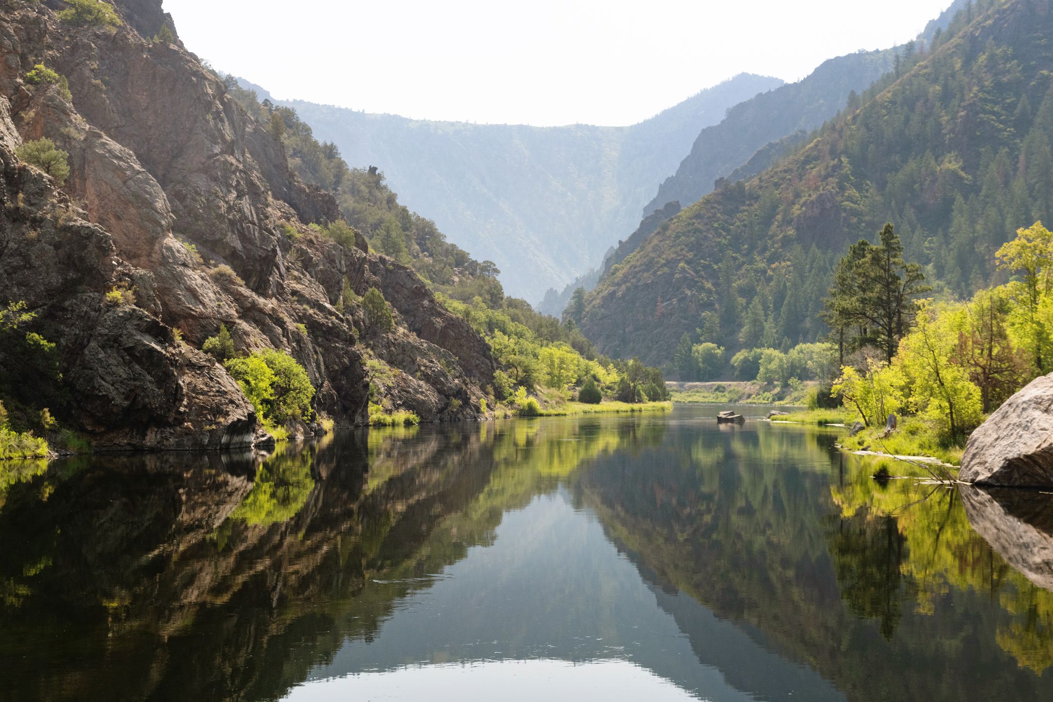 The tall mountains covered with trees reflect in the river waters at the East Portal of Black Canyon of the Gunnison national park in Colorado, USA on a hazy summer day.