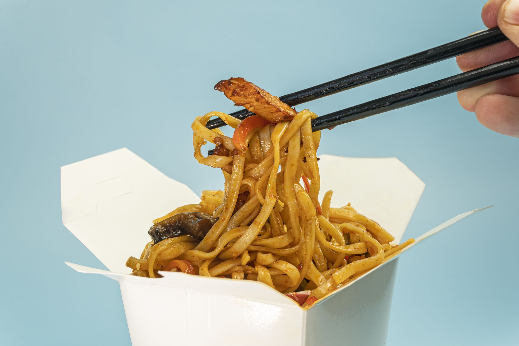Udon stir-fry noodles with chicken meat and sesame in cardboard box on blue background close up. bright blue background color.