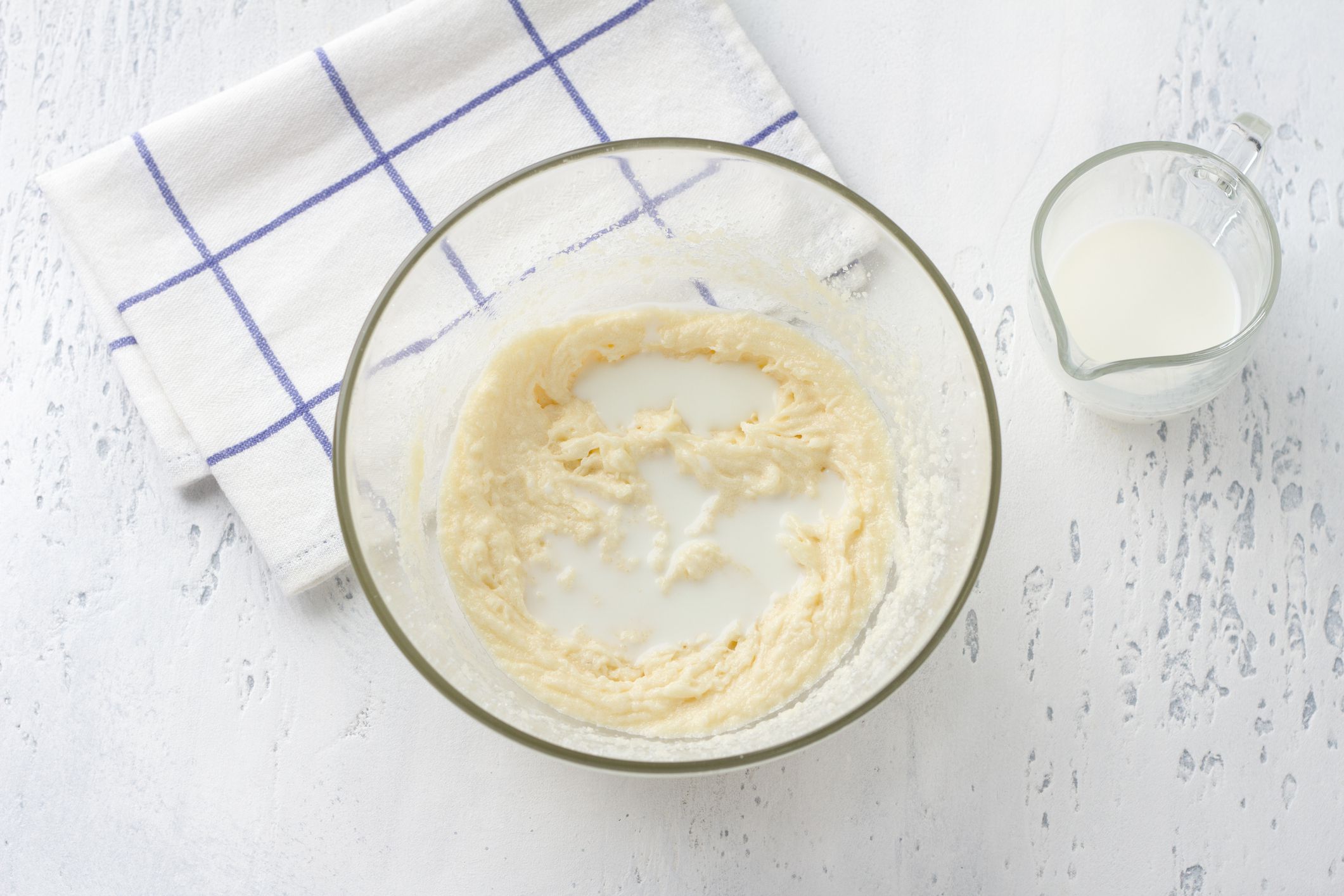 Glass bowl with whipped egg-cream-sugar mass, adding milk on a light blue background, top view. Cooking homemade milk cakes, do it yourself, step by step, step 4