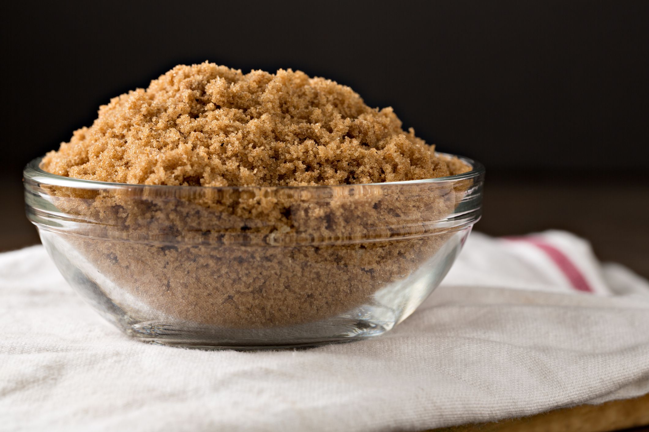 An extreme close up, isolated horizontal photograph of a glass bowl full of brown sugar sitting on a white tea towel.