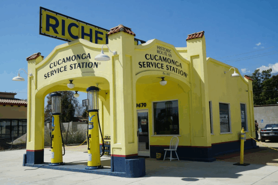 A bright yellow historic gas station with &ldquo;Cucamonga Service Station&rdquo; signs, vintage gas pumps, and a large &ldquo;RICHFIELD&rdquo; sign on the roof, under a clear blue sky.