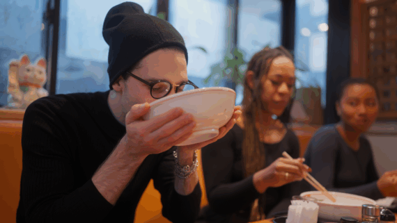 A person in a beanie and glasses sips from a bowl at a restaurant, while two others beside them eat with chopsticks. The scene is casual, with a bright interior and a lucky cat figurine in the background.