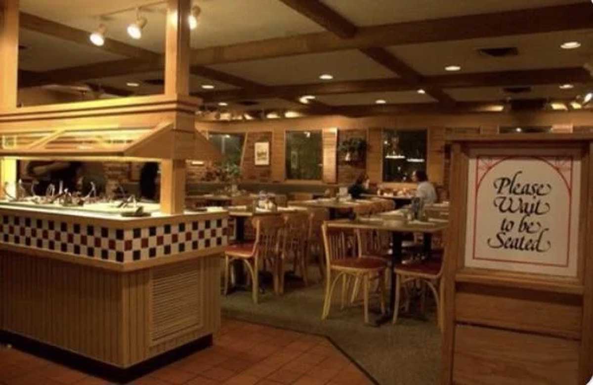 A cozy restaurant interior with wooden tables and chairs, an empty salad bar on the left, and a sign by the entrance that says, "Please wait to be seated." The scene evokes classic Vintage Pizza Hut photos with a few customers seated in the background.