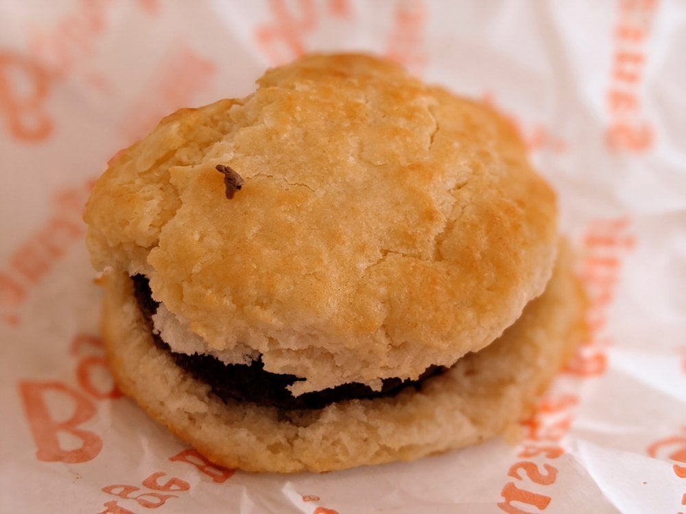 A close-up of a sausage biscuit on branded paper. The biscuit is golden brown and flaky, with a sausage patty visible between the biscuit halves.