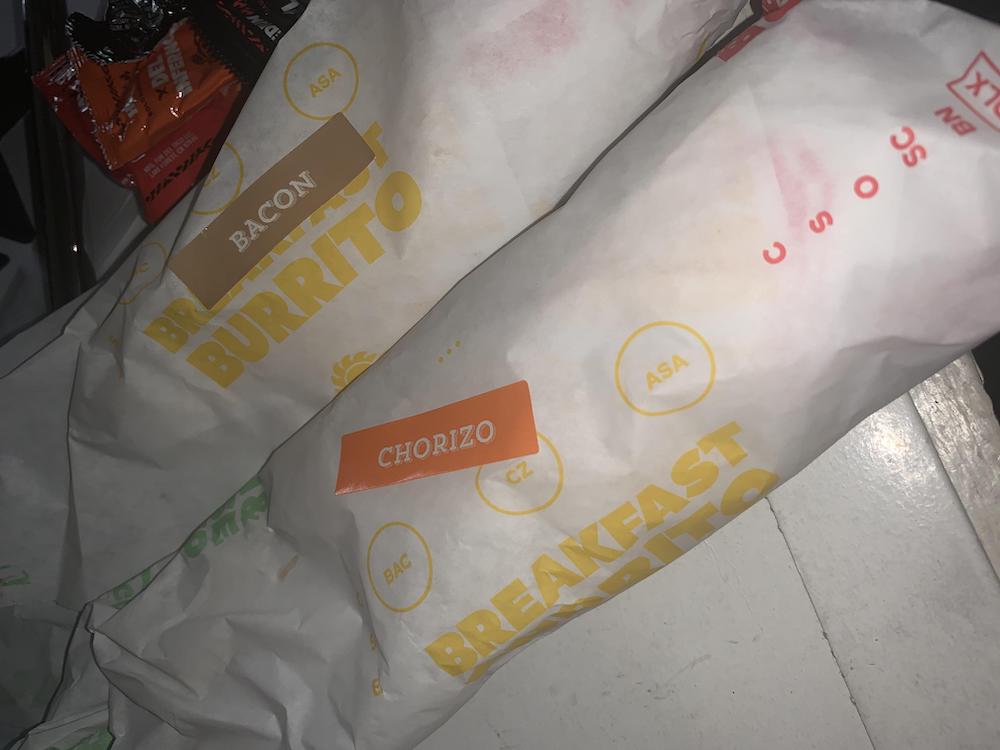 Two wrapped breakfast burritos labeled "Bacon" and "Chorizo" are placed side by side on a white surface. Packets of sauce are visible in the background.