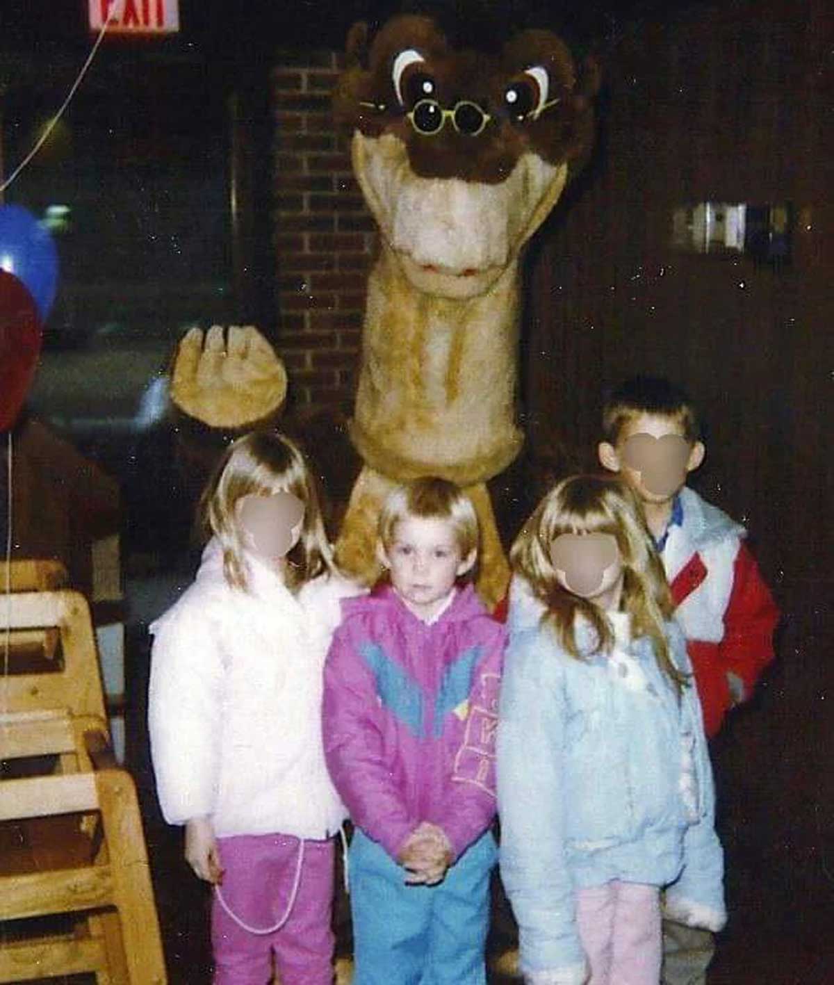 Four children pose indoors in front of a large, friendly camel mascot, reminiscent of Vintage Pizza Hut photos. The kids wear winter jackets, their faces blurred for privacy. Balloons and brick walls create a festive backdrop.
