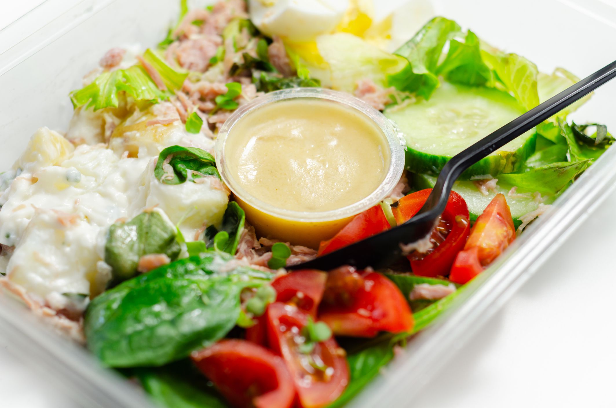Healthy salad in the plastic food box, potato salad with hard boiled egg, shredded ham, cucumber, tomatoes, lettuce and spinach with a honey and mustard dressing pot, takeaway meal