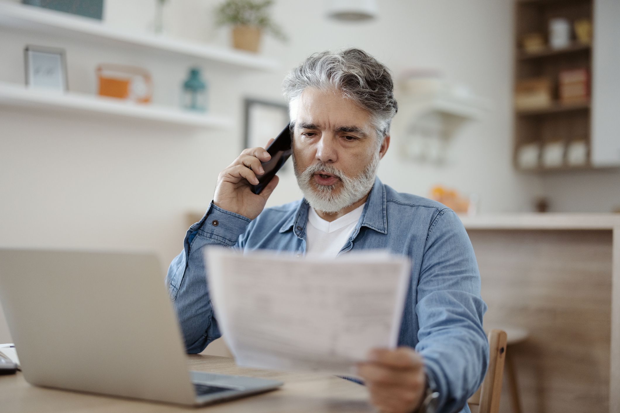 Discussing document. Mature man holding paper and talking on phone at home