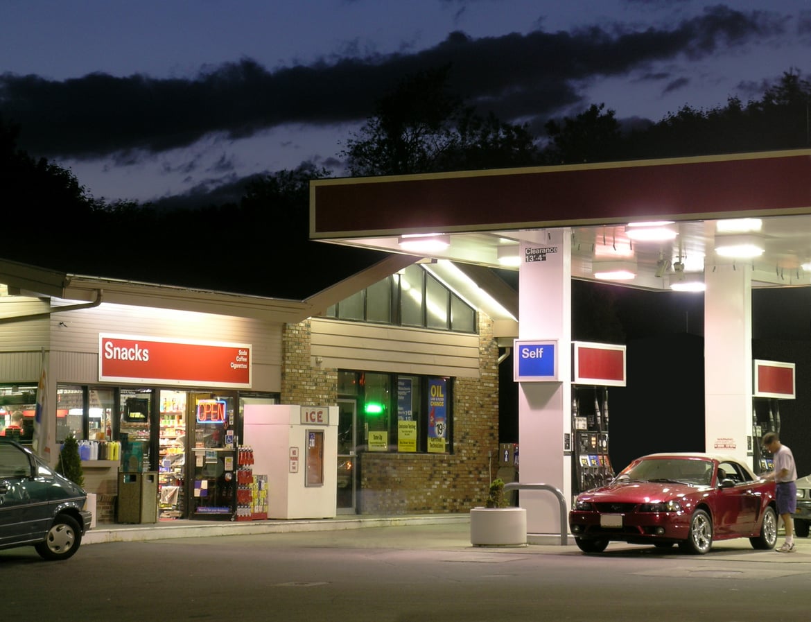 A red sports car is parked at a self-service gas pump at night, with a person refueling. The brightly lit convenience store labeled "Snacks" and "ICE" is visible in the background.
