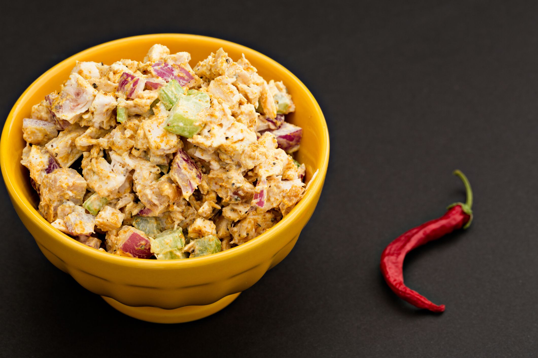 A high angle close up shot of a yellow bowl full of chicken curry salad and a red cayenne pepper.