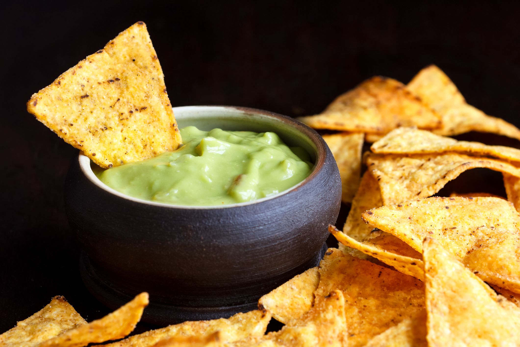 Round rustic brown bowl of guacamole dip surrounded with tortilla chips in perspective. Black background.