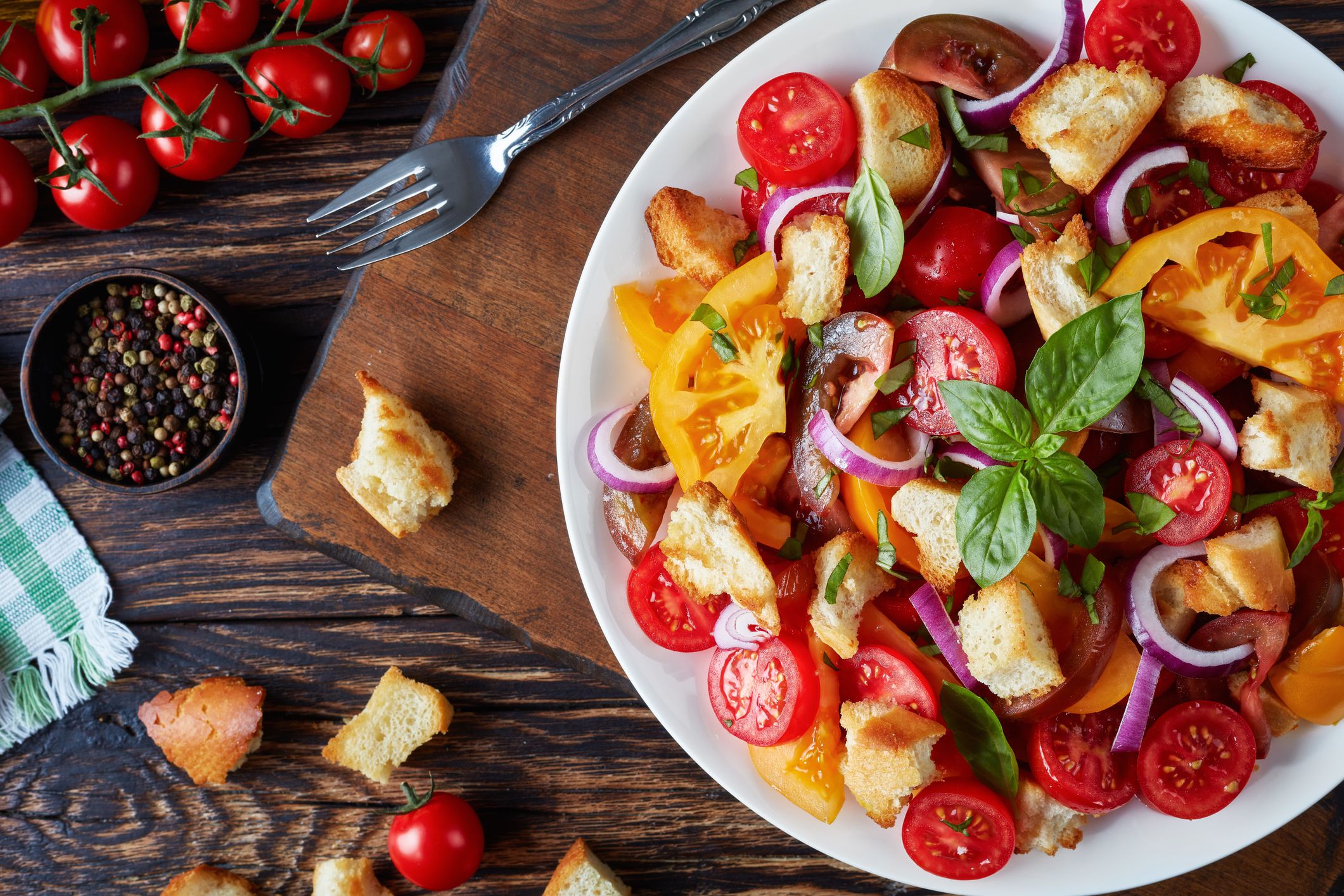 delicious fresh classic italian salad panzanella with tomatoes, croutons basil and onion rings on a white plate on an old wooden table with ingredients, view from above, flat lay, close-up