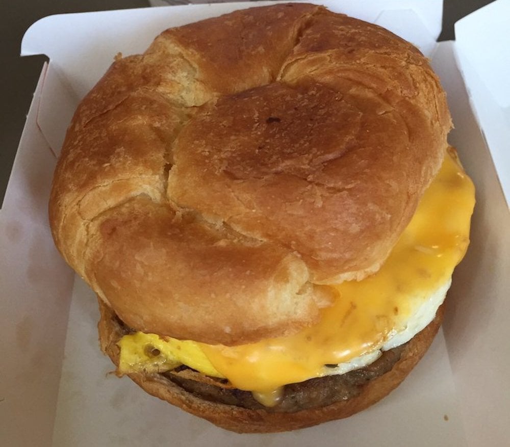 A breakfast sandwich with a fluffy croissant bun, a slice of melted cheddar cheese, cooked egg, and sausage, served in a white takeout box.