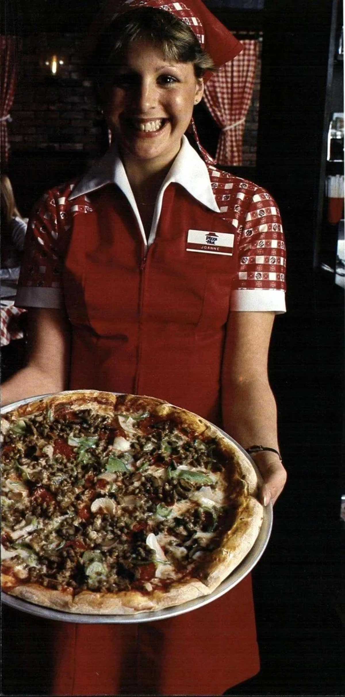 A smiling waitress in a red and white uniform holds a large pizza topped with meats and vegetables, standing in a restaurant with checkered curtains—a scene reminiscent of Vintage Pizza Hut photos.