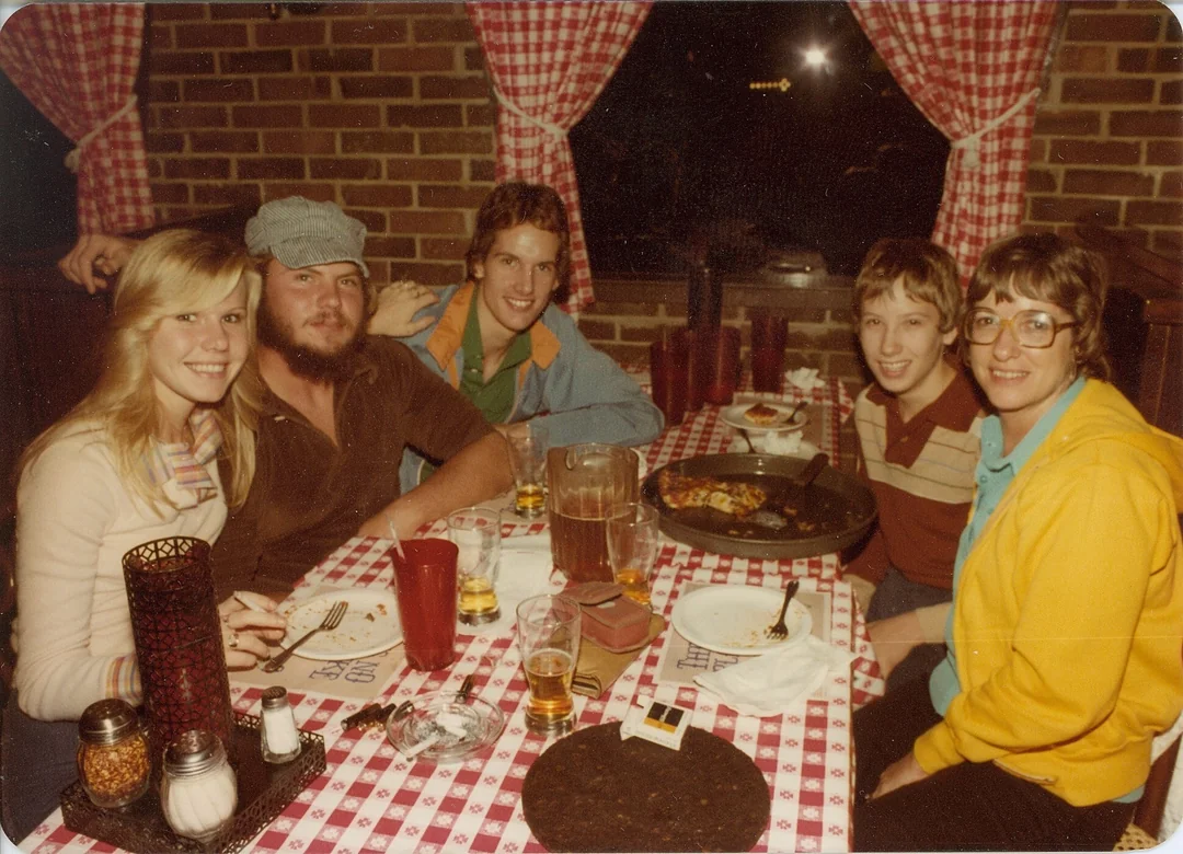 Five people sit around a table with a red checkered tablecloth in a cozy restaurant, smiling at the camera. There are drinks, a pizza, and plates on the table. Brick walls and checkered curtains are in the background.