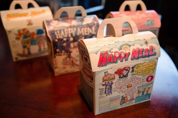 Four colorful McDonald&rsquo;s Happy Meal boxes with playful cartoon designs are displayed on a wooden table, with one box in sharp focus at the front and three more slightly blurred in the background.