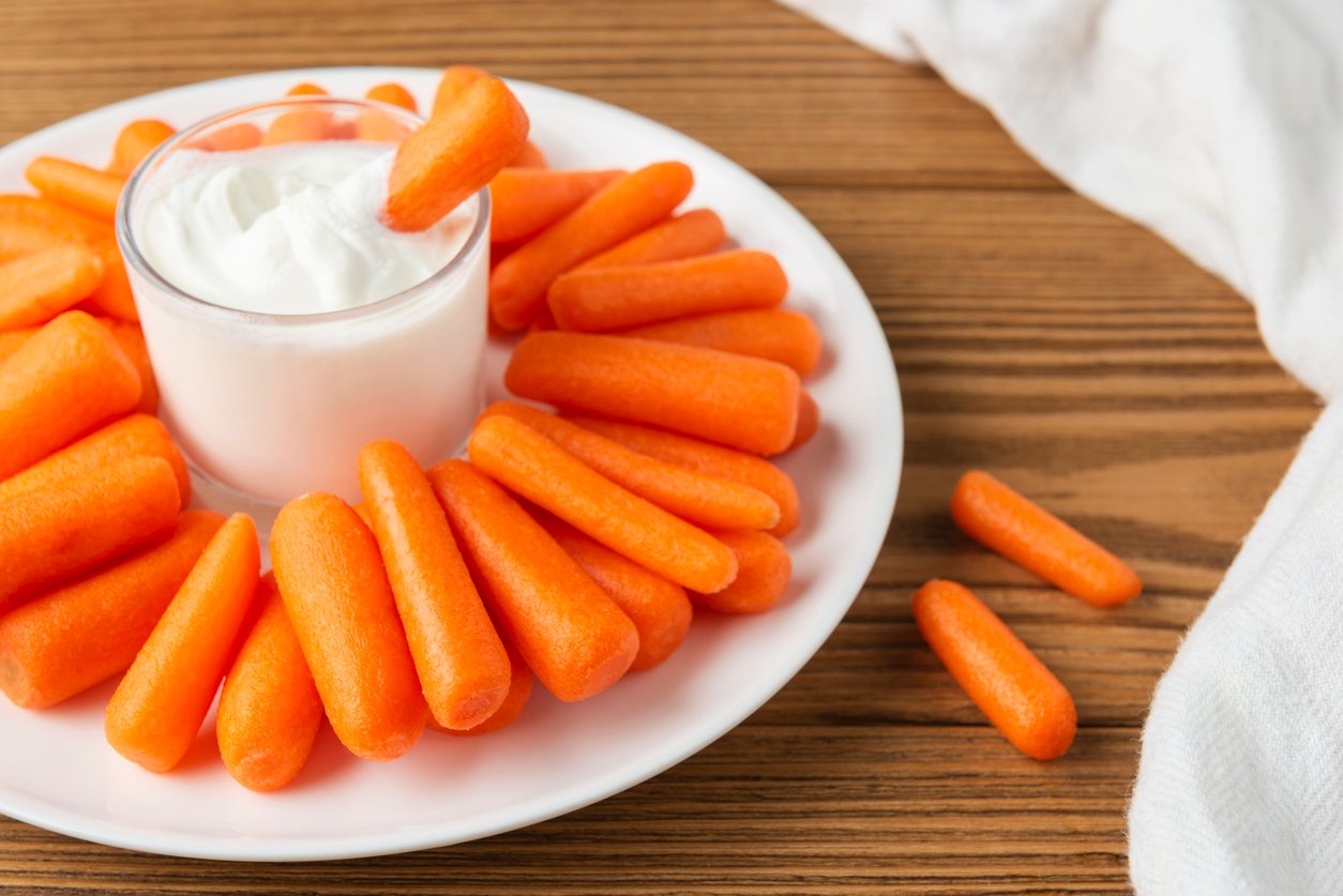 A white plate with fresh baby carrots, a classic grocery store purchase, arranged in a circle around a glass cup filled with white dip, sits on a wooden table with a white cloth nearby.