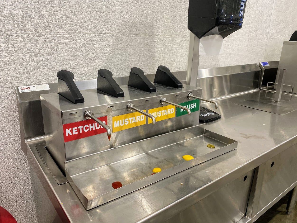 Condiments area for the food court at Costco, four pumps for ketchup, two mustards ones, and relish, and napkins, clean stainless steel table, white wall
