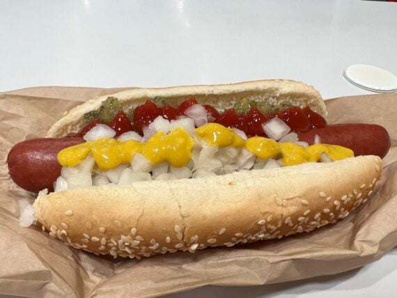 A sesame seed hot dog bun with a sausage topped with chopped onions, yellow mustard, relish, and ketchup, just like a classic Costco hot dog, resting on brown paper.
