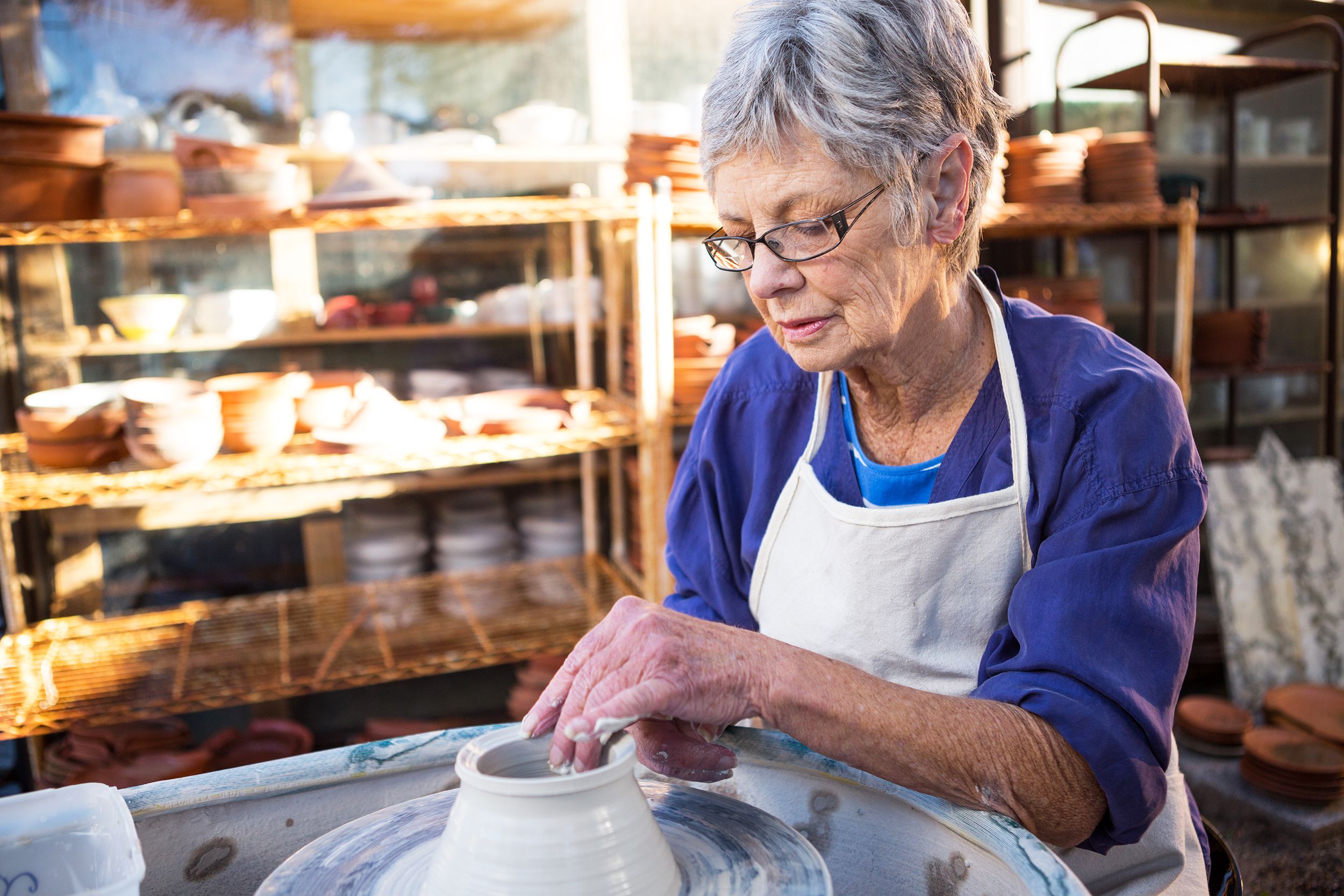 Focus on an attentive senior woman at a pottery wheel, making a vase with clay in a pottery workshop with sunlight coming inside, rows of different kinds of pottery blurred in the background
