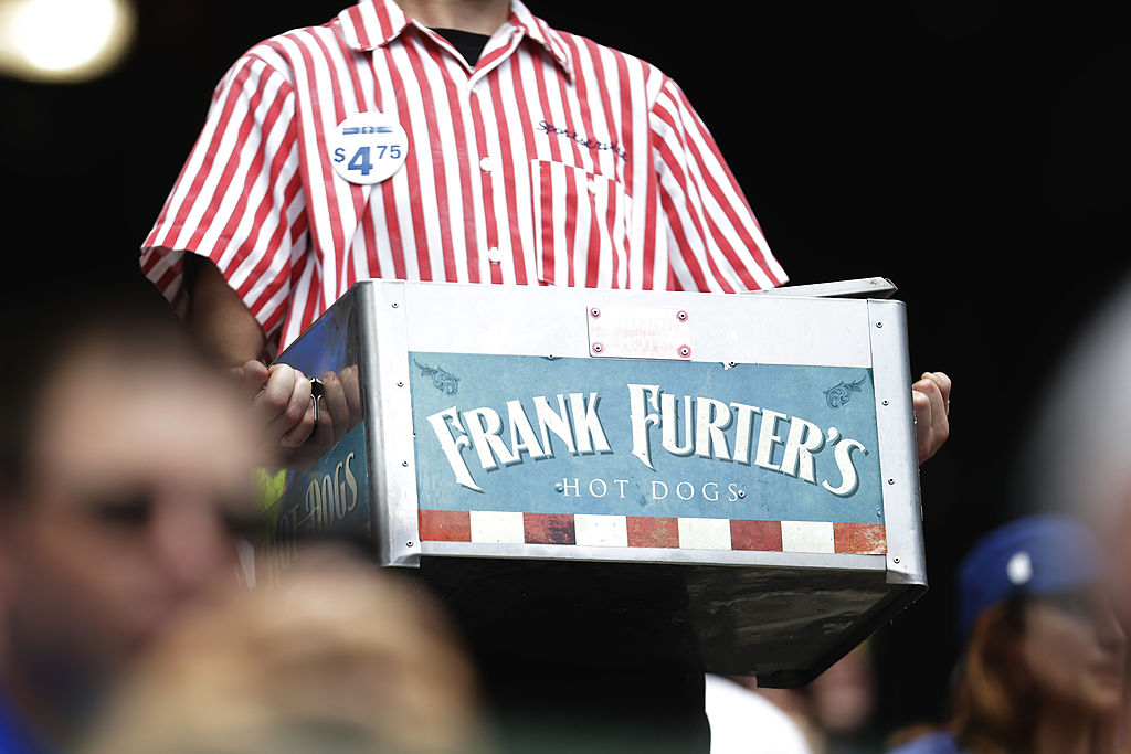 A vendor in a red-and-white striped shirt holds a metal box labeled "Frank Furter’s Hot Dogs" with a price button showing $4.75—nearly matching the famous Costco hot dog price—selling hot dogs at an event or stadium.