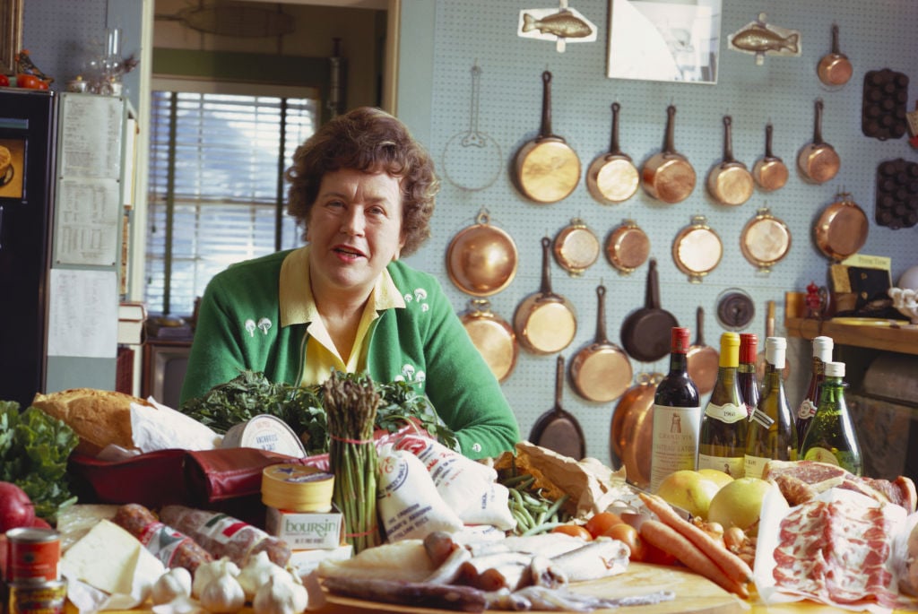 A woman with short brown hair sits at a kitchen table covered with fresh ingredients and bottles, perhaps planning a meal inspired by the famous Costco hot dog. Behind her, copper pans hang in her bright, cozy kitchen.